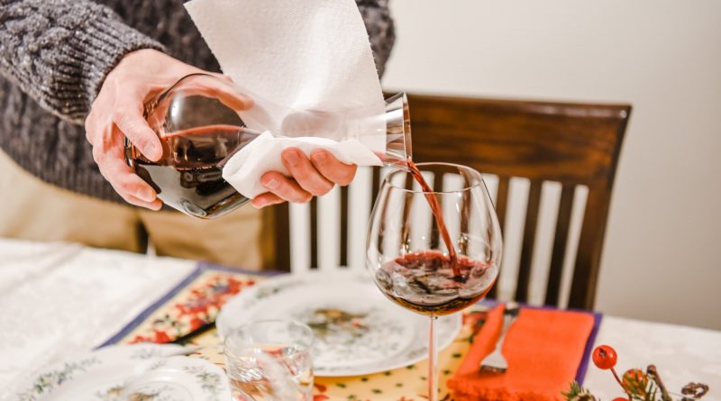 A man pours red wine into a wine glass prior to dinner guests taking a seat at the table.