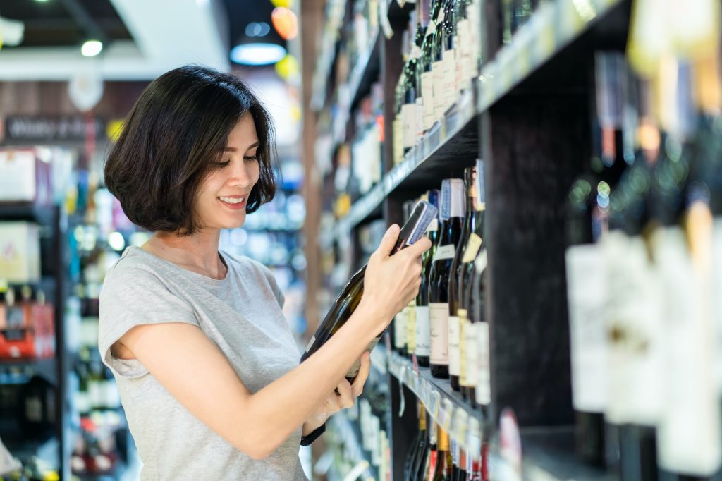 Asian beautiful woman choosing nice red wine bottle picking up from alcohol shelf in supermarket