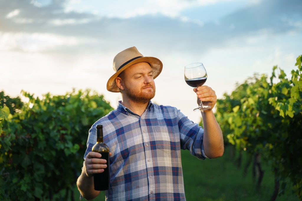 Millennial vintner holding a glass and a bottle of organic bio red wine outdoors in a vineyard.