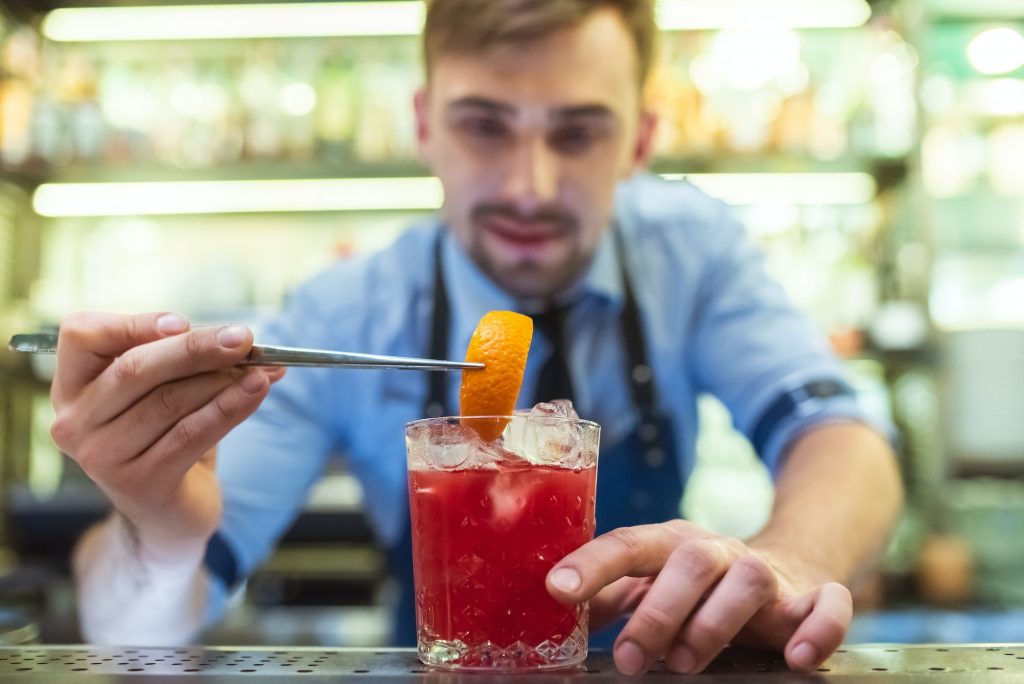 The bartender making a red cocktail with an orange on the bar