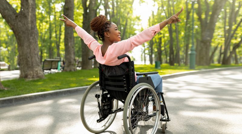 Handicapped black woman in wheelchair on walk at city park, showing victory gesture, feeling