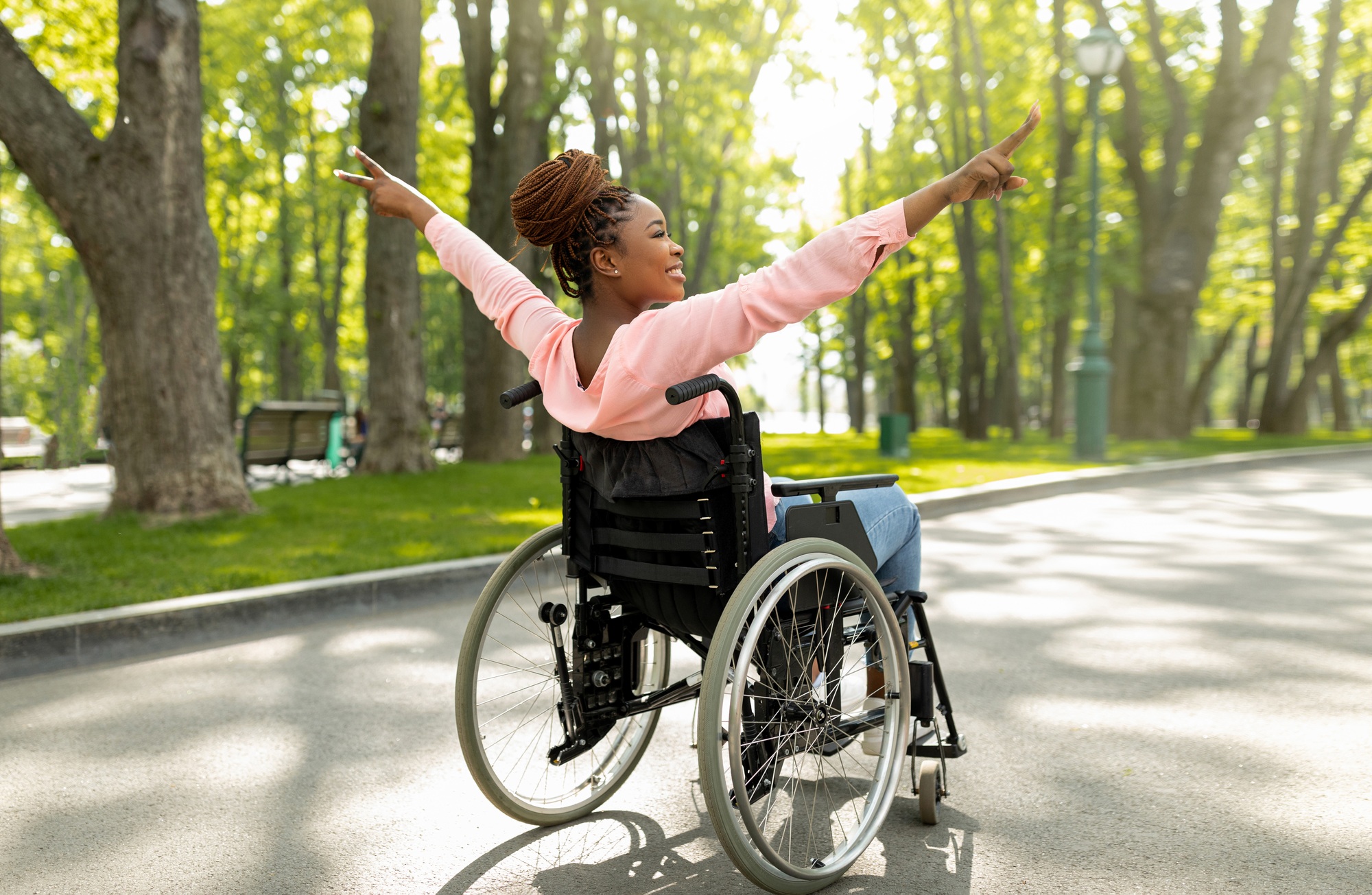 Handicapped black woman in wheelchair on walk at city park, showing victory gesture, feeling